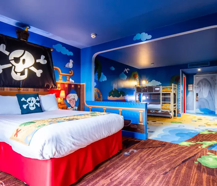 A pirate themed room, a double bed with a jolly roger above it is in the forground with a treasure chest themed bunk bed in the background