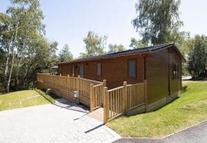 Wooden cabin with fenced deck in sunny, tree-lined area.