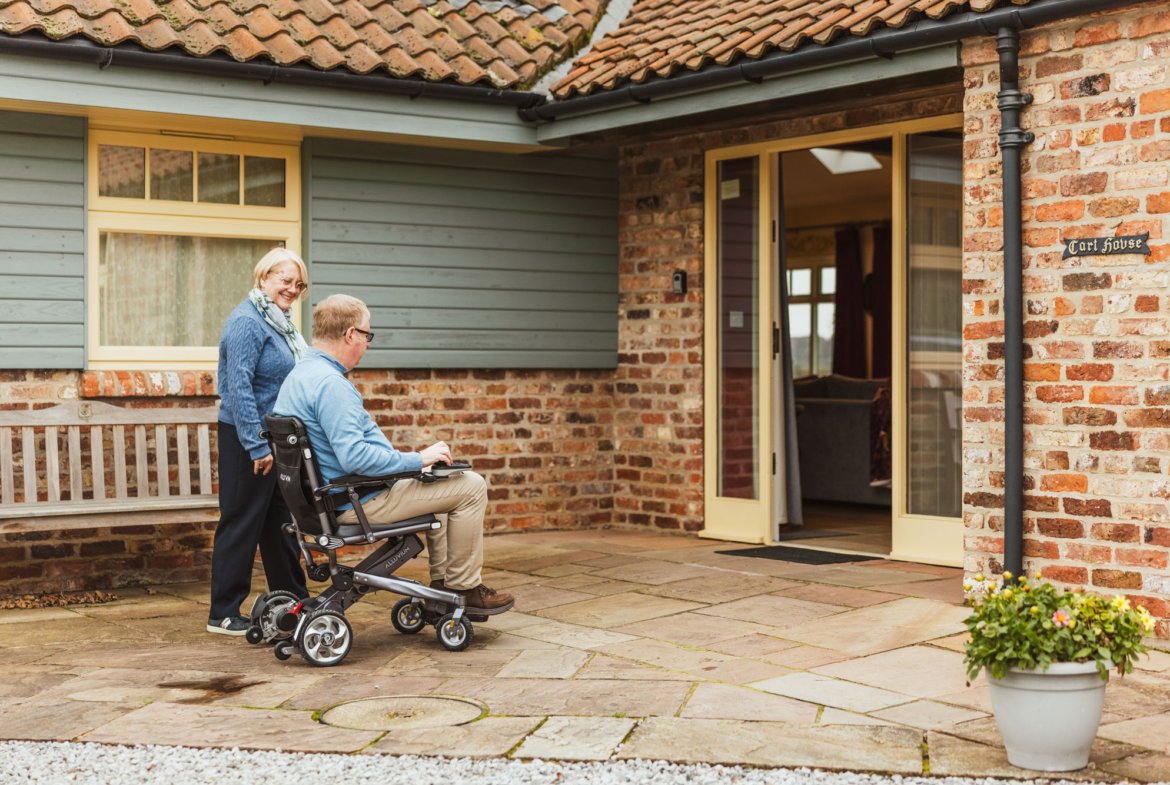 Woman assists man in wheelchair entering house.