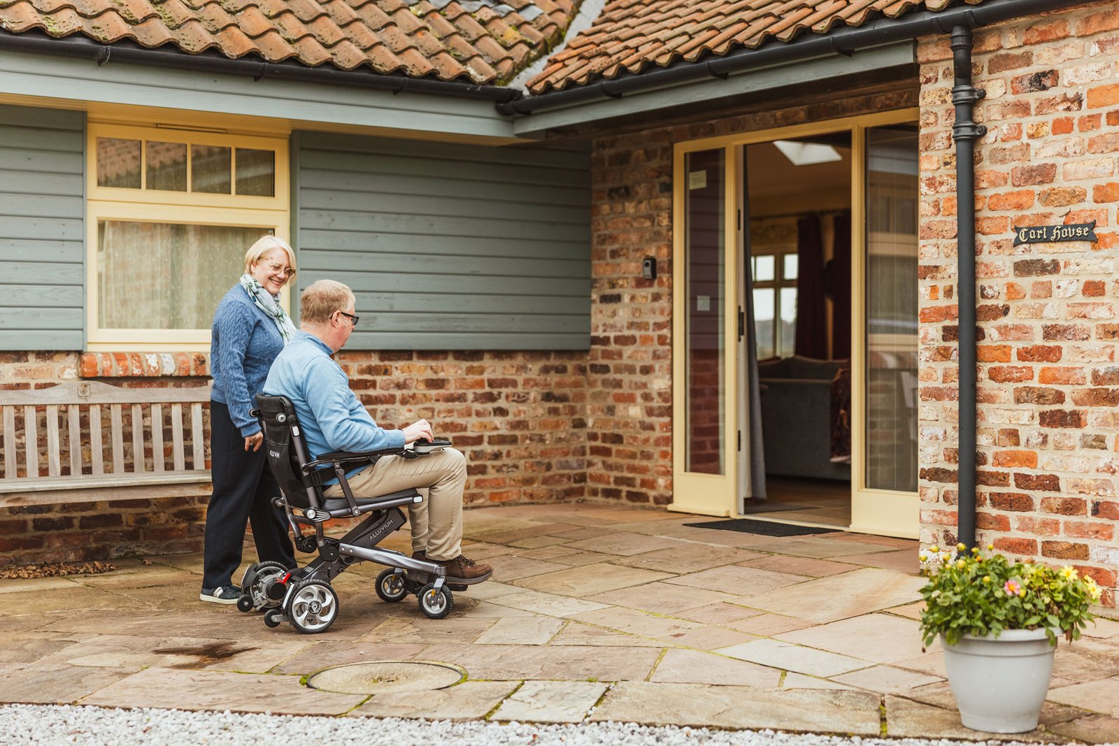Woman assists man in wheelchair entering house.