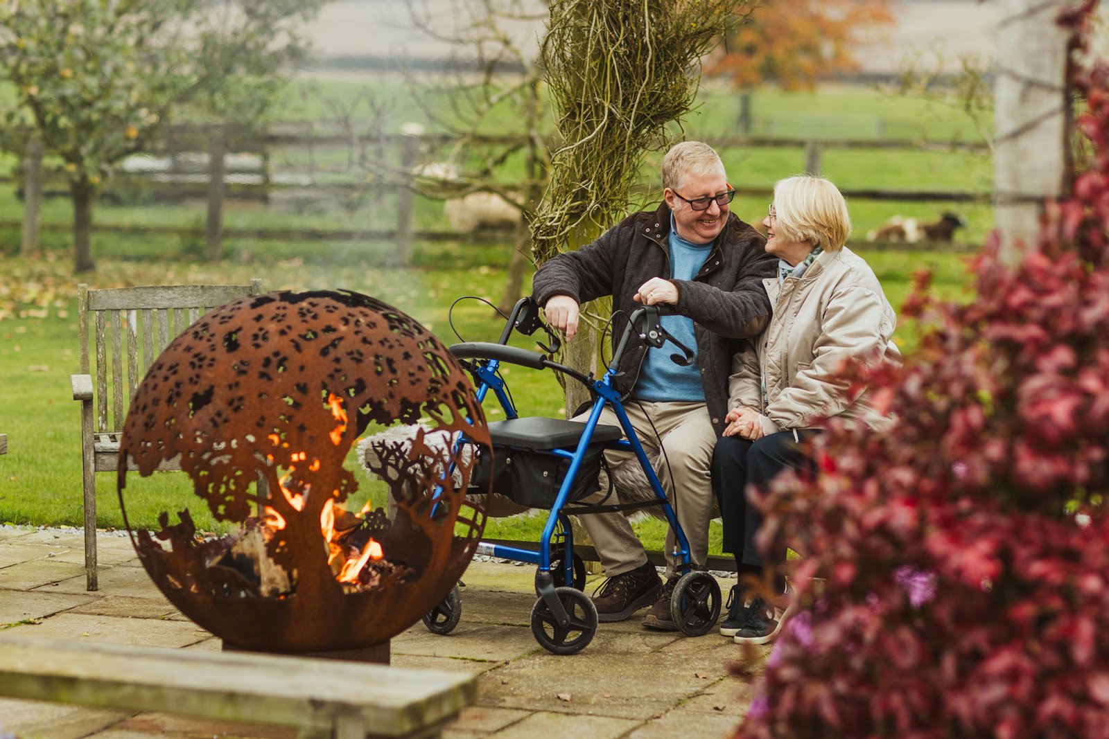 Elderly couple sitting by outdoor fire, smiling.