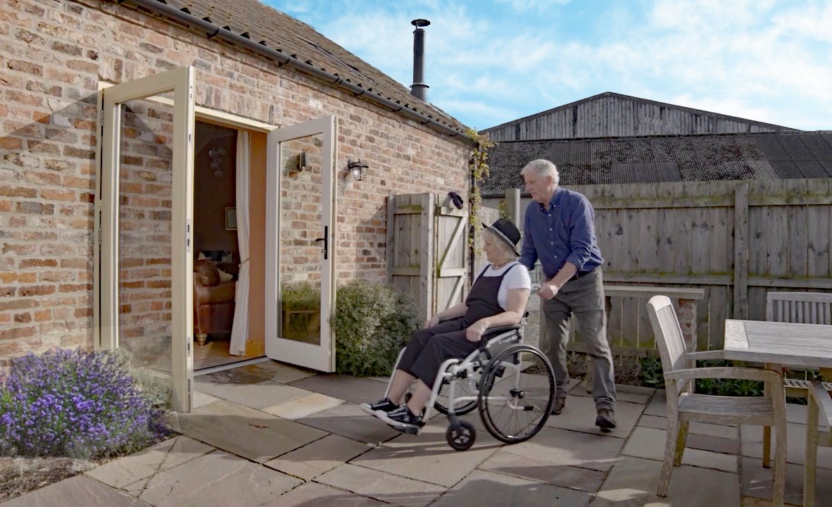 Man pushes woman in wheelchair outside cottage